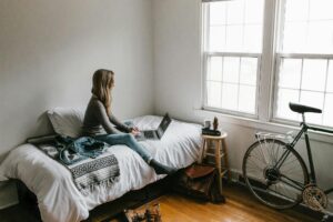 A young woman works on a laptop in a cozy, minimalist bedroom setting, showcasing remote work lifestyle.